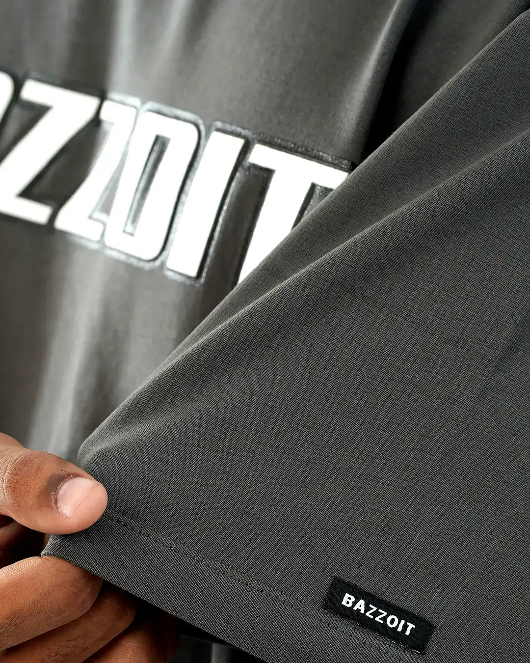 Gray t shirt with 'BAZZOIT' branding held by a hand, showing close-up of fabric texture.