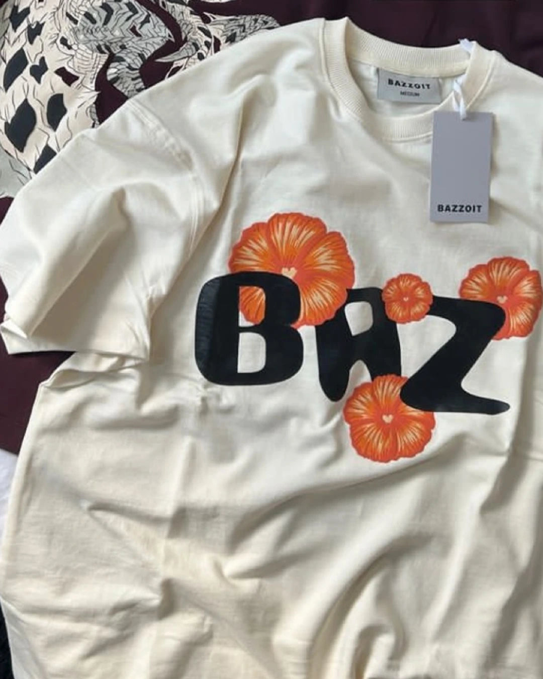 White t-shirt with 'Baz' text and orange flowers, displayed on a surface.